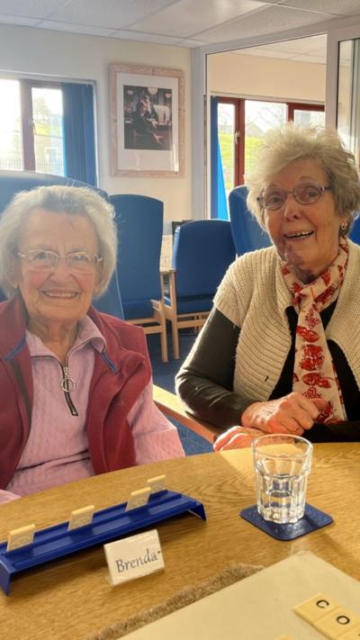 Two women sat a table in a day care centre. One is wearing a pink top with dark pink sleeveless jacket. The second woman has a black top, white jacket and red and pink scarf. Both women are wearing glasses
