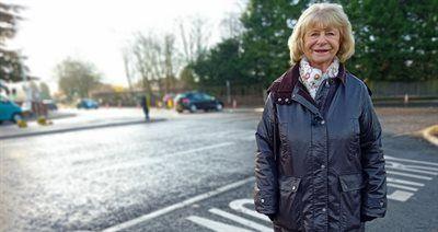 A woman with blonde hair to her shoulders stands next to a road. There are trees behind her and some cars parked across the road.