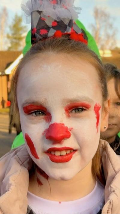 Evelyn is a schoolgirl and has her dark blonde hair pulled up on top of her head, she has white and red clown make up and is wearing a puffy pink coat over her costume. To her right is a child with a red wig and a brown mask wearing a pair of "bloodstained" dungarees.