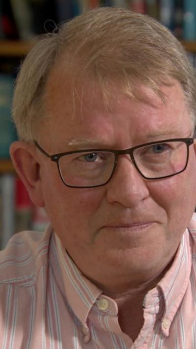 Dr Kieran McManus in pink shirt with black glasses, he has short grey hair
