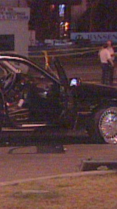 A black vehicle with an open passenger door sits on a road during nighttime. There are officers and caution tape in the background.