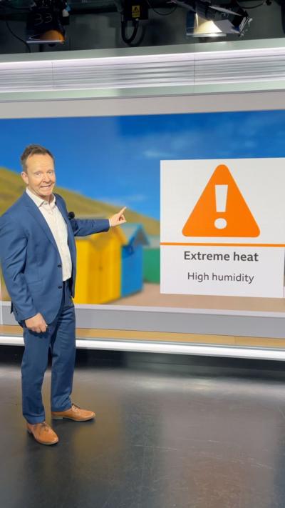 Man in a blue suit stands in a TV studio pointing to a Extreme Heat sign