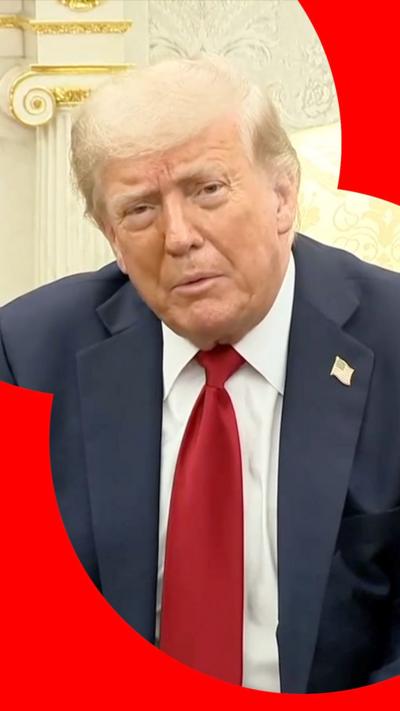 US President Donald Trump looking straight into the camera, whilst sitting in the Oval Office. He's wearing a navy blue suit, with a pin of US flag on it and a red tie. 