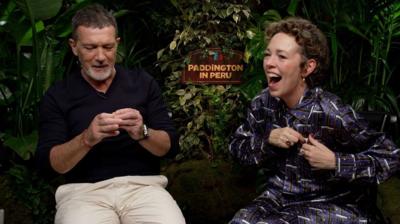 Antonio Banderas looking at a Blue Peter badge next to Olivia Colman who is excitedly putting her badge on. A sign in the background reads Paddington in Peru