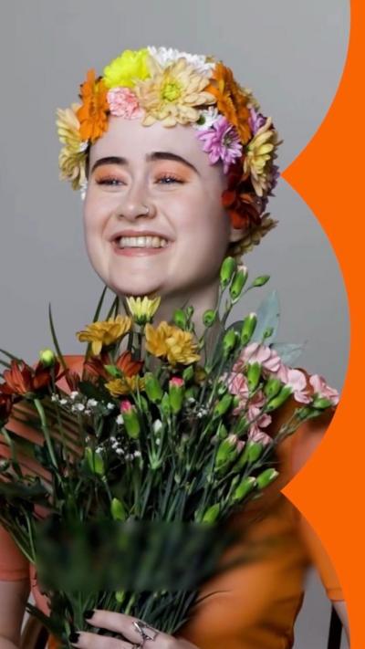 A young woman smiling, wearing a crown of flowers, orange eyeshadow and holding a bunch of carnations.