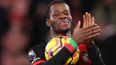 A smiling Dango Outtara wears red and black Bournemouth kit as he applauds with hands held high and a yellow football tucked under his right arm