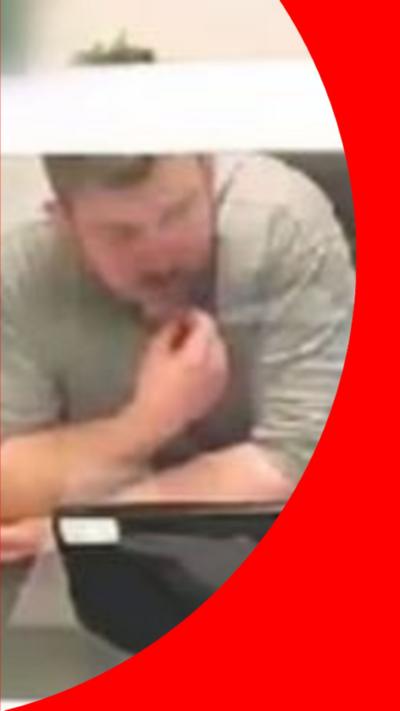McCullagh in a grey tshirt behind a glass screen looking at a TV screen. 