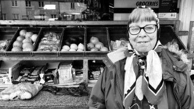 A woman in a headscarf and a winter jacket is standing in front of a stall selling fruit and vegetables.