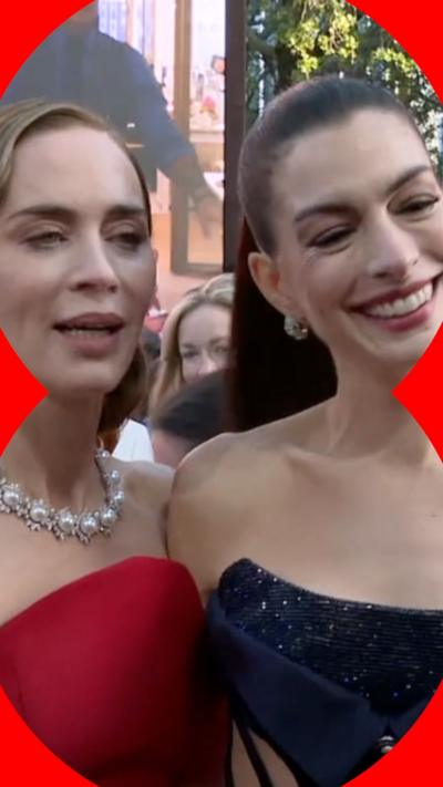 Emily Blunt and Anne Hathaway laugh and talk to an interviewer, while holding each other in a sideways hug.