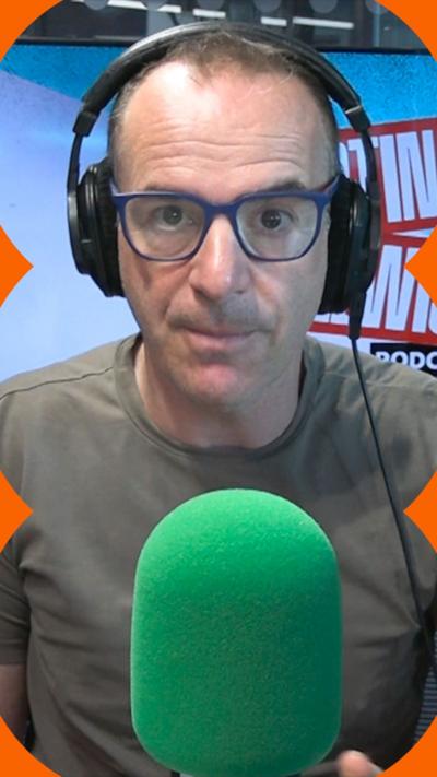 Martin Lewis is wearing dark green T-shirt and black-framed glasses. He is wearing headphones and sitting behind a green mic.