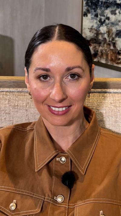 A woman with dark hair tied back is looking into the camera. She has dark eyes and red lipstick. She is wearing a light brown to with white buttons and is sitting on a beige seat.
