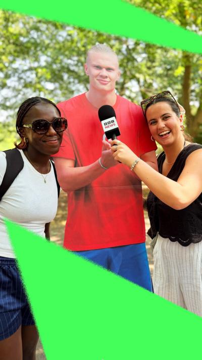 Two young women pose in front of a life-size cutout of Erling Haaland in a park.