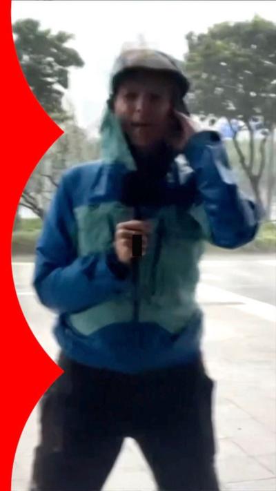 Laura Bicker in a blue raincoat with the hood up, she's holding a microphone and being blown by the wind