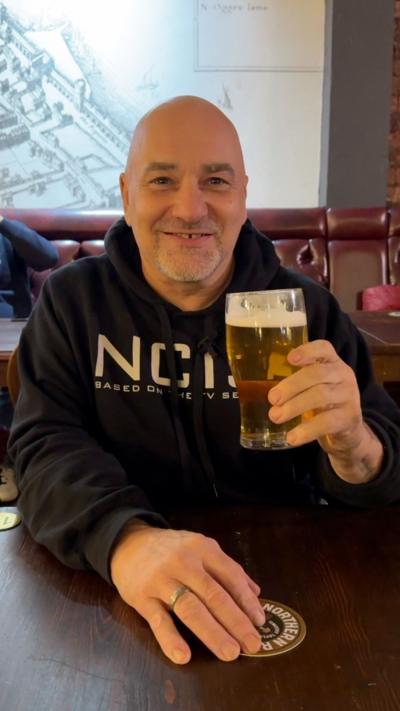 Simon Berry sits at a wooden table in a traditional pub, wearing a dark hoodie and holding a pint of pale beer. The cosy setting features leather bench seating, wooden floors, brick walls, and nearby patrons.