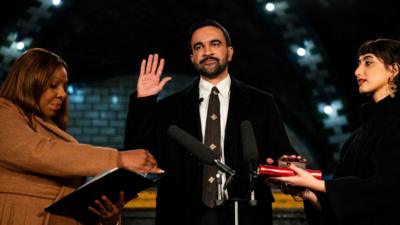Zohran Mamdani holds his right hand in the air as he takes an oath.