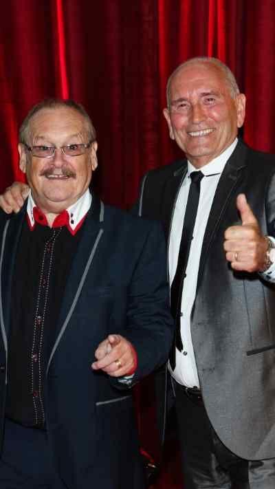 A man with a moustache and glasses on the left is with a taller, thinner bald man on the right. Both are dressed in suits and smiling.