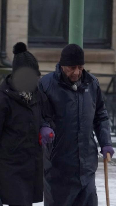 A man in a dark blue jack and hat walks along using a walking stick. He is holding the arm of a person in a black bobble hat and coat whose face has been blurred out.