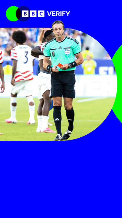 A football referee on a pitch holds a red card. Behind him footballers walk on a pitch. 