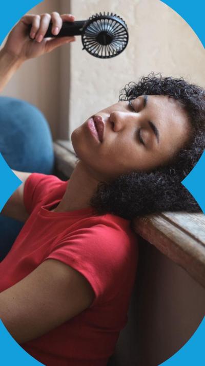 Woman in red top sits against a wall with a handheld fan. She has her eyes closed and her head rested on a windowsill. 