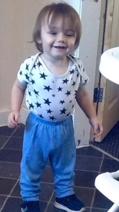 A two-year-old boy standing up wearing a vest with black stars and blue jogging bottoms