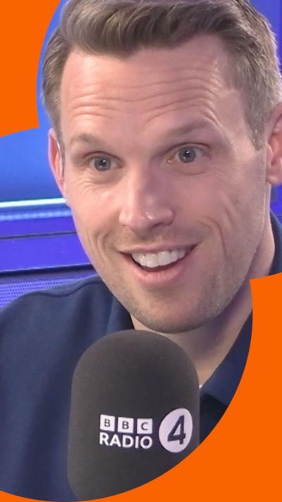 Greg Foot smiles as he talks into a BBC Radio 4 microphone. He wears a blue shirt. 