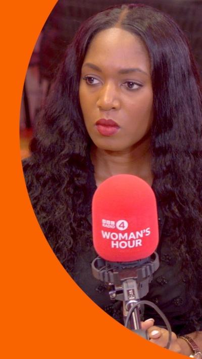 A woman with very long dark brown wavy hair sits behind a microphone. She is wearing red lipstick and a dark-coloured top.