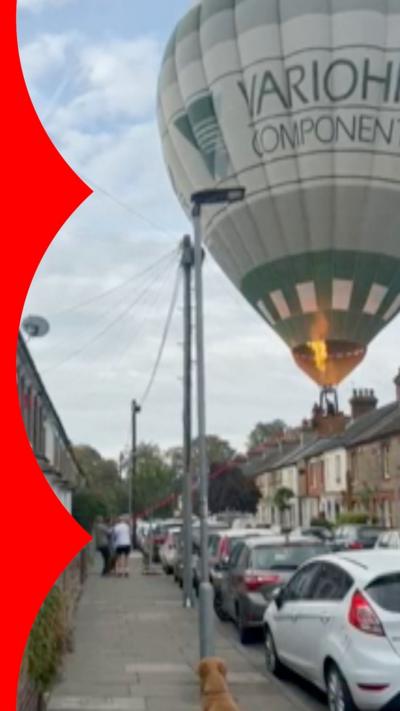 A hot air balloon lands in the middle of a residential street in Bedford. The balloon can be seen coming down between terrace houses, landing in between two rows of parked cars - the thumbnail shows the balloon with fire being blown into the balloon