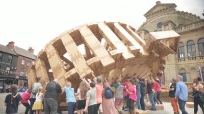 Group of people lifting up a large cardboard structure of a wheel with a triangle point at the top.