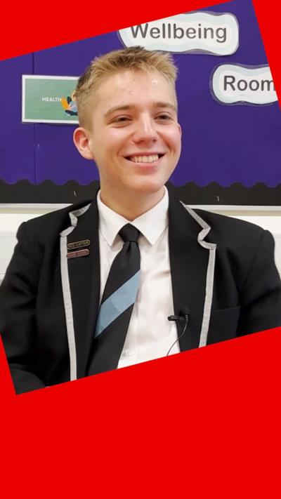 A student smiles - he has short blond hair and he wears a dark blazer jacket, white shirt and a dark tie with a light blue stripe