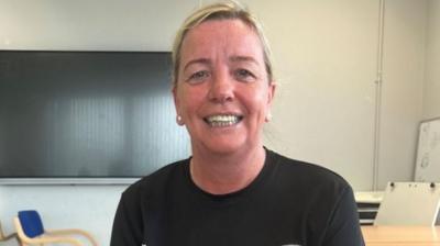 A woman with tied up blond hair smiles towards the camera. She is wearing a black t-shirt. 