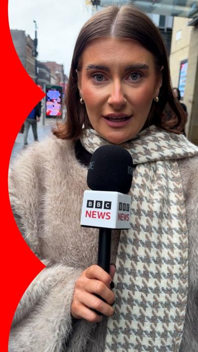 Woman looking at camera. She is wearing a cream and light grey geometric print scarf, has short brown hair and is wearing gold small hoop earrings.