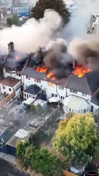 Aerial view of a residential building fire, with flames breaking through the roof of multiple attached houses and dense smoke spreading across the area