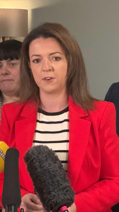 Liz Kimmins is speaking at a presser with microphones infront of her. She has long brown hair and wearing a red jacket with a black and white striped top underneath.
