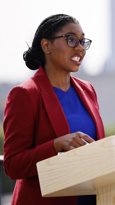 Kemi Badenoch speaking at a podium in a maroon jacket and blue top