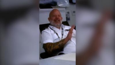 A police officer in white shirt, sitting back in a chair and smiling. He's bald with a beard.
