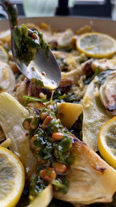 A close-up of a spoon drizzling pine nuts and greens over baked fennel and lemon slices.