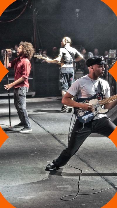 A man on a stage sings into a microphone. Two other men with guitars are on stage with him