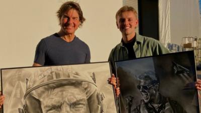 Actor Tom Cruise standing next to artist Wilf Elliott. They are outside and standing in front of a white wall, both holding framed charcoal portraits of Cruise.
