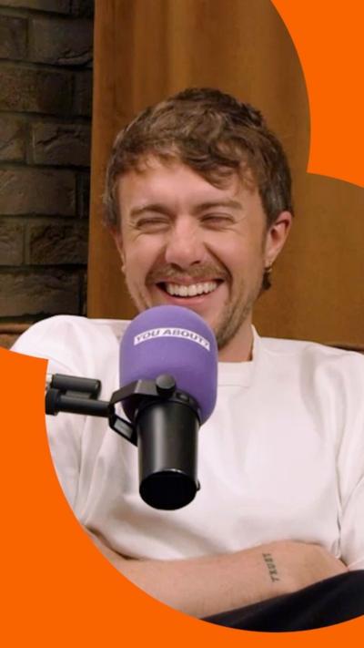 A man, Roman Kemp, in a white T-shirt laughs with his arms crossed behind a mic