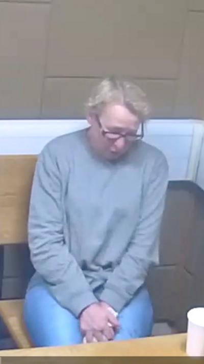 Two women are sat down in a room on wooden chairs. The woman on the right of the image has her face blurred and is wearing a grey cardigan with a white stripe on the sleeves and mid-section. The woman on the left is wearing a grey jumper and glasses. The room is cream coloured with square and rectangular shapes in the wall.
