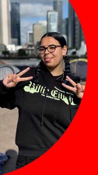 A woman wearing a Billie Eilish hoodie strikes a pose. She is standing in a makeshift campsite, with tents behind her.