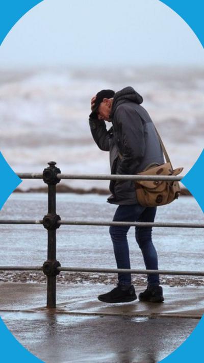 A man clutches his hat while walking along a windy seafront with rough waves in the background