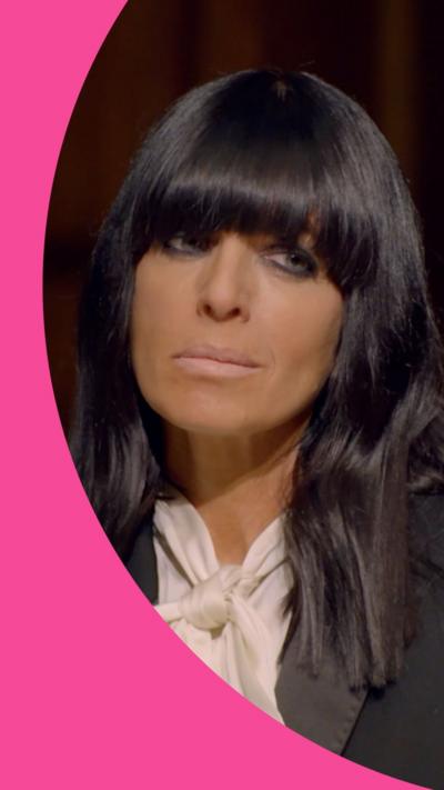 Claudia Winkleman looks mischievously to the side. She has long black hair with a fringe, and wears a black suit jacket
