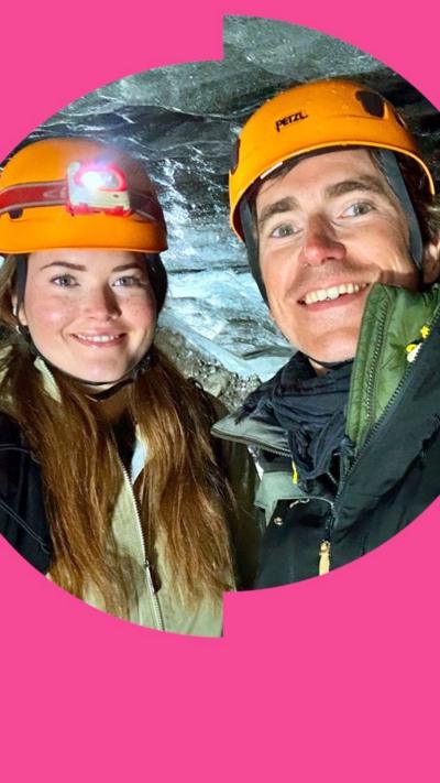 A woman and a man smile inside an ice cave - they both wear orange helmets and winter jackets