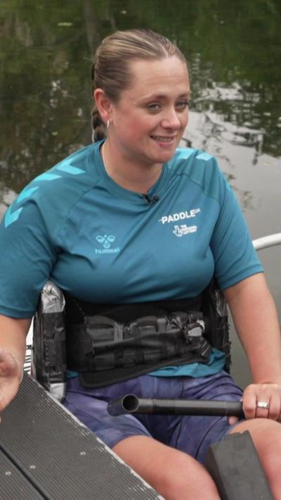 A woman with tied back blonde hair sat in a kayak on a riverside. She is wearing a turqoise top with purple shorts
