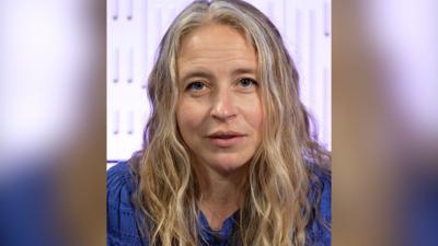 A woman with long blond wavy hair wearing a blue top, looks at the camera