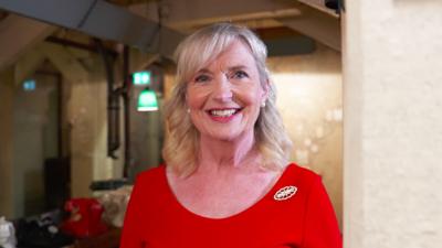 Carol Kirkwood, wearing a red dress and brooch, smiling in the map room of Churchill's War Rooms
