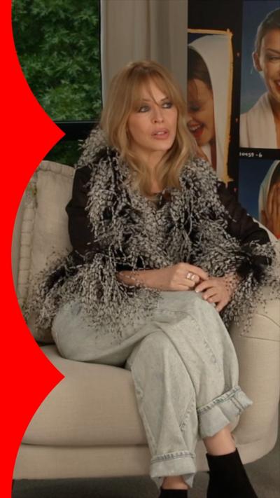 Kylie Minogue has long blonde hair and is wearing a fluffy black and grey top, and grey trousers. 