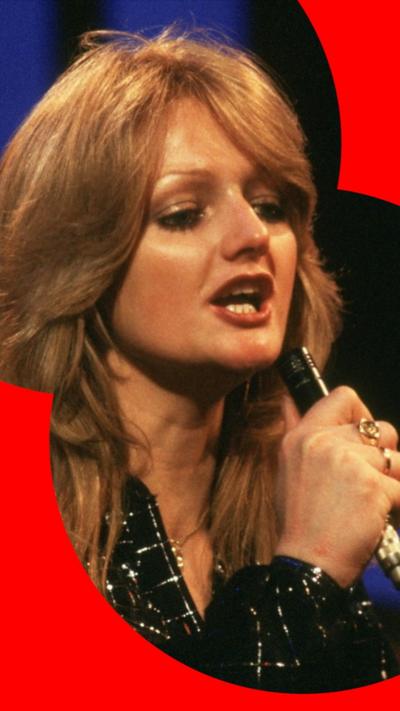 Bonnie Tyler wth long brown hair and singing into a microphone, and wearing a sparkly black suit.