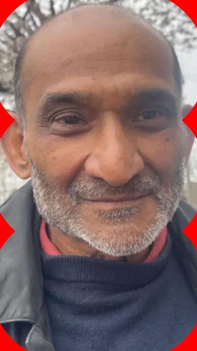 A man with a short grey beard is smiling. He is wearing a shirt with a red collar, a blue jumper and a black jacket.